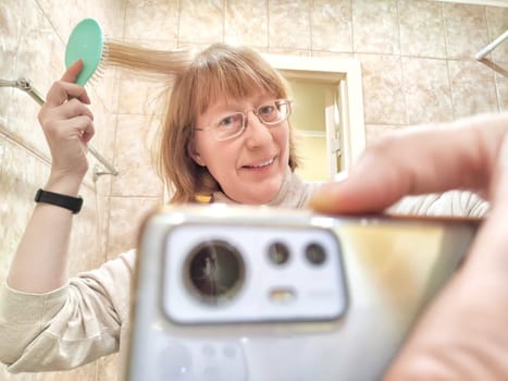 Shaggy, funny blonde middle-aged woman in glasses doing hair and taking Blogger girl posing for blog, stream