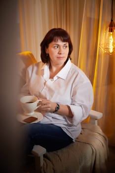 Nice girl with a mug of tea or coffee in cozy room. A middle-aged adult woman relaxing in the evening in the living room or bedroom in the evening