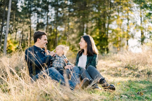 Mom looks at dad touching little daughter nose with his finger while sitting in a clearing. High quality photo