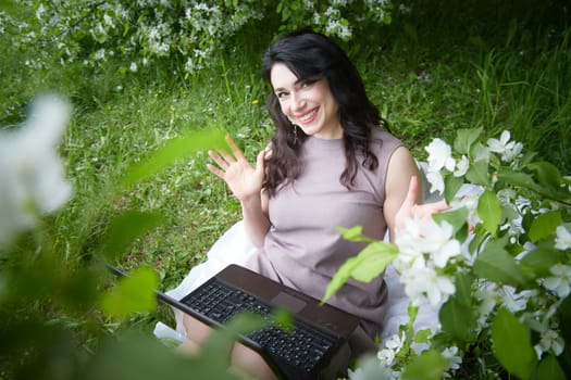 Girl engages with laptop amidst lush greenery. Freelancer Woman Working on Laptop Outdoors in a Blooming Garden