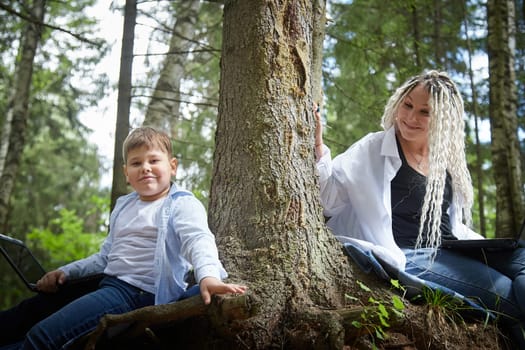 Mother and son with a laptops in the forest in summer. Fat young smart teenage boy and woman working with modern IT technologies in nature