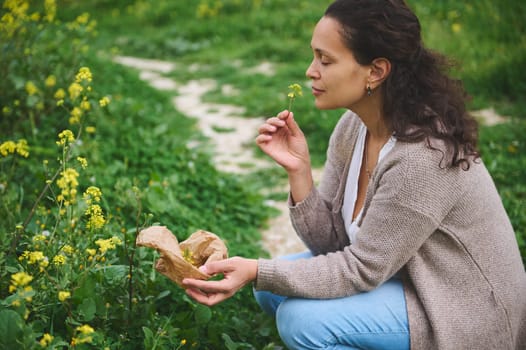 Beautiful young woman herbalist sniffing a flower while collecting medicinal herbs in the mountains outdoors, preparing healing remedy and tea according to traditional recipe. Alternative medicine