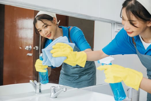 In rubber gloves a cheerful maid wipes bathroom mirror with a rag ensuring cleanliness and shining reflection. Hotel service emphasizes professional cleaning for hygiene. Maid cleaning at home