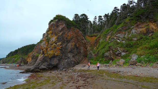 People walk on shore with rocks and trees. Clip. People are walking on beautiful beach with large rocky block. Walk on northern beach with rocks on cloudy summer day.