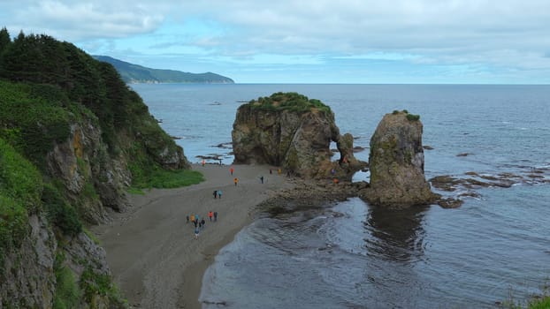 People on shore with rocks and arch. Clip. Group of people walks on northern coast of sea with rocky arches. Top view of people walking on rocky shore.
