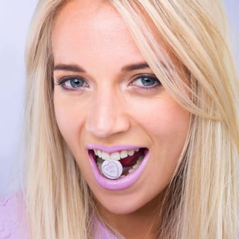 Woman, beauty and studio portrait with candy in mouth with makeup, lipstick or heart sign by purple background. Girl, person and model in closeup with sweets, emoji or happy for eating with cosmetics.