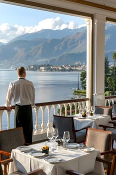 A man on a balcony enjoying the scenic view of a lake with mountains in the background. The property is furnished with tableware and furniture, creating a lovely interior design