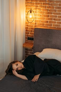 A woman is lying on a bed in a bedroom, looking relaxed and thoughtful while surrounded by cozy furnishings. Vertical frame.