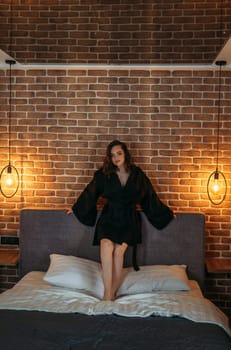 A woman is seated on a bed placed against a brick wall, creating a simple and intimate setting. Vertical frame.