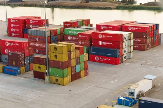 Seville, Spain - June 2, 2023: Multiple colorful shipping containers are neatly stacked at a port terminal, with cranes visible in the background, suggesting active logistics operations.