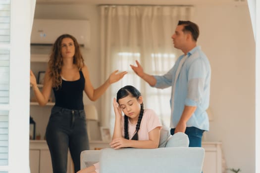 Annoyed and unhappy young girl sitting on sofa trapped in middle of tension by her parent argument in living room. Unhealthy domestic lifestyle and traumatic childhood develop to depression.Synchronos