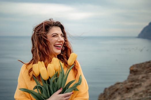 Portrait of a happy woman with hair flying in the wind against the backdrop of mountains and sea. Holding a bouquet of yellow tulips in her hands, wearing a yellow sweater.