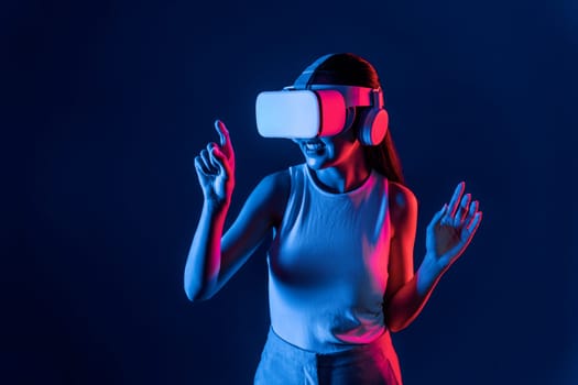 Smart female stand with surrounded by cyberpunk neon light wear VR headset connecting metaverse, futuristic cyberspace community technology. Woman using finger pointing virtual object. Hallucination.