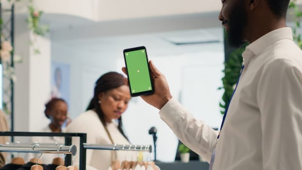 African american man holding mockup cellphone in premium clothing store with stylish formalwear clothes. BIPOC customer with green screen mobile phone in designer showroom