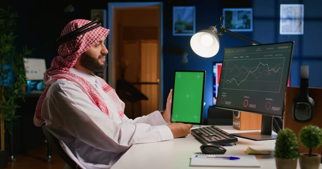 Muslim worker in apartment office holds chroma key tablet, crosschecking data on it with business report graphs on computer. Smiling entrepreneur with mockup digital device in hand performs job tasks