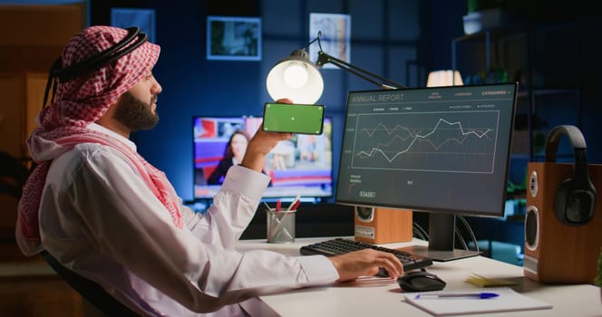 Arab man watching video on green screen cellphone while looking at business annual report charts on computer. Worker spending time on chroma key mobile phone while solving job tasks at home