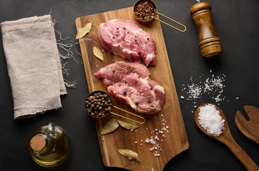 Two raw pork neck steaks on a board and spices for cooking. Top view of black table. Copy space