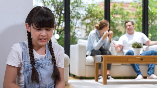 Stressed and unhappy young girl huddle in corner, cover her ears blocking sound of her parent arguing in background. Domestic violence at home and traumatic childhood develop to depression. Synchronos