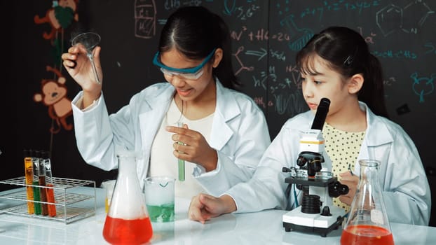 Young highschool student doing experiment by pouring sample in test tube. Girl wearing glasses and looking under microscope to inspect sample in STEM science class. Creative education. Edification.