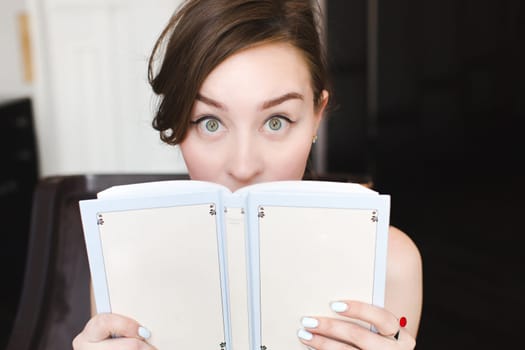 Woman hiding behind the grey book. Woman covering her face. Woman hiding face behind book looking at camera surprise. Education concept. Face expression. Girl holding book.