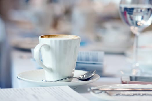 Simple white porcelain cup full of hot aromatic coffee placed on saucer with teaspoon on blurred background of served table