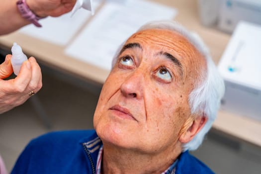 Close-up top view of a senior man looking up while ophthalmologist applying drops in the eyes in a clinic