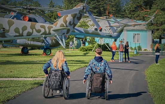 Two individuals, a man and a woman, both in wheelchairs, are exploring an outdoor museum featuring retired military helicopters on a sunny day, showcasing accessibility and inclusion.