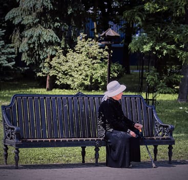 An elderly woman wearing a black dress and white hat sits on a park bench, surrounded by lush trees. The serene expression on her face suggests that she is enjoying the peace and quiet of the park.