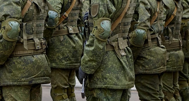 Army soldiers in uniform stand in formation on a concrete surface with yellow crosswalk lines. They wear combat fatigues and boots, showing discipline and readiness.