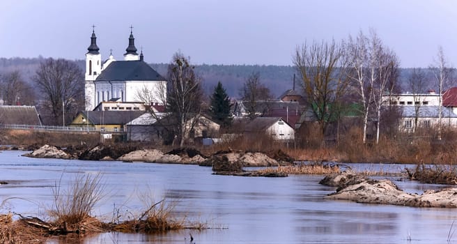 A picturesque European village by a serene river, surrounded by greenery and colorful flowers. White church, quaint houses, blue sky with fluffy clouds - a perfect setting for a peaceful spring day.