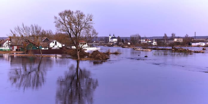 A picturesque European village by a serene river, surrounded by greenery and colorful flowers. White church, quaint houses, blue sky with fluffy clouds - a perfect setting for a peaceful spring day.