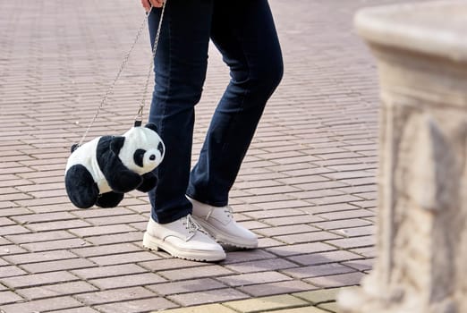A fashionable young woman strolls with a cute panda plush bag, donning a chic ensemble and white kicks. The panda accessory brings a fun, whimsical touch to her urban style.