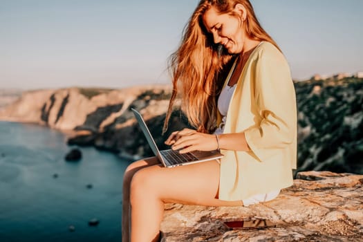 Successful business woman in yellow hat working on laptop by the sea. Pretty lady typing on computer at summer day outdoors. Freelance, travel and holidays concept.