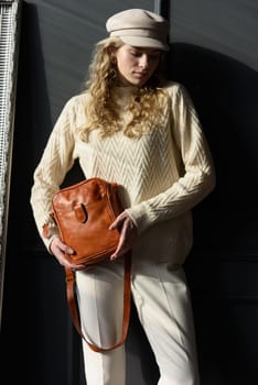 Studio portrait of beautiful woman with a curly blond hair holding brown bag, posing on gray background. Model wearing stylish cap, sweater and classic trousers