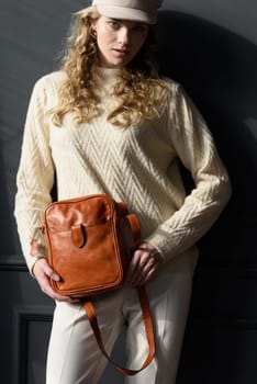 Studio portrait of beautiful woman with a curly blond hair holding brown bag, posing on gray background. Model wearing stylish cap, sweater and classic trousers