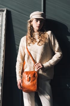 Studio portrait of beautiful woman with a curly blond hair holding brown bag, posing on gray background. Model wearing stylish cap, sweater and classic trousers