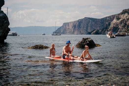 Father and his adorable little son and daughter sitting on stand up board having fun during summer beach vacation