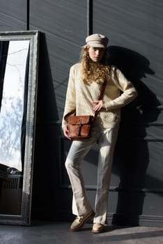 Studio portrait of beautiful woman with a curly blond hair holding brown bag, posing on gray background. Model wearing stylish cap, sweater and classic trousers