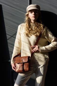 Studio portrait of beautiful woman with a curly blond hair holding brown bag, posing on gray background. Model wearing stylish cap, sweater and classic trousers