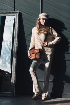 Studio portrait of beautiful woman with a curly blond hair holding brown bag, posing on gray background. Model wearing stylish cap, sweater and classic trousers