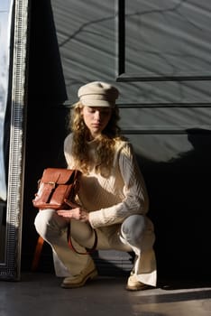 Studio portrait of beautiful woman with a curly blond hair holding brown bag, posing on gray background. Model wearing stylish cap, sweater and classic trousers