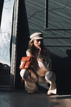 Studio portrait of beautiful woman with a curly blond hair holding brown bag, posing on gray background. Model wearing stylish cap, sweater and classic trousers