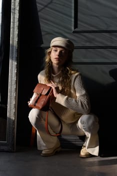 Studio portrait of beautiful woman with a curly blond hair holding brown bag, posing on gray background. Model wearing stylish cap, sweater and classic trousers