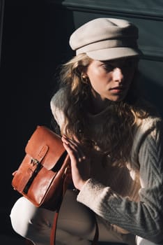 Studio portrait of beautiful woman with a curly blond hair holding brown bag, posing on gray background. Model wearing stylish cap, sweater and classic trousers