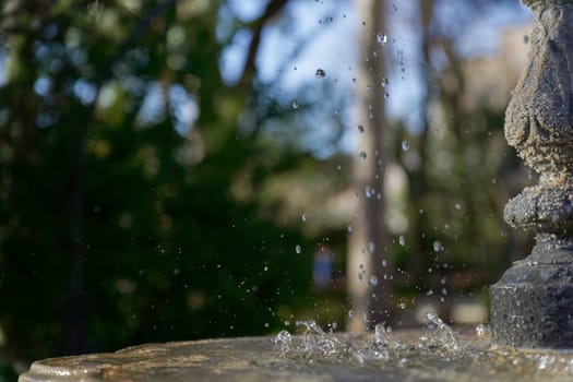 profile view of a public fountain with splashing water, blue sky and park trees in the background.