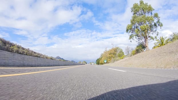 On a crisp winter day, a car cruises along the iconic Highway 101 near San Luis Obispo, California. The surrounding landscape is brownish and subdued, with rolling hills and patches of coastal vegetation flanking the winding road.