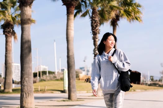 young asian woman dressed in sportswear smiling happy while walking with yoga mat hanging on her arm, sport and healthy lifestyle concept