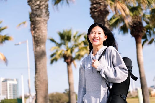 young asian woman dressed in sportswear smiling happy while walking with yoga mat hanging on her arm, sport and healthy lifestyle concept