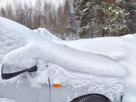 A car is almost entirely blanketed by a thick layer of fresh snow, with only a portion of the side mirror and front wheel visible. Heavy snowfall and cold weather conditions