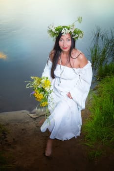 Adult mature brunette woman in a white dress, sundress and a wreath of flowers in summer by the water of river or lake in evening at sunset. Celebration of the Slavic pagan holiday of Ivan Kupala